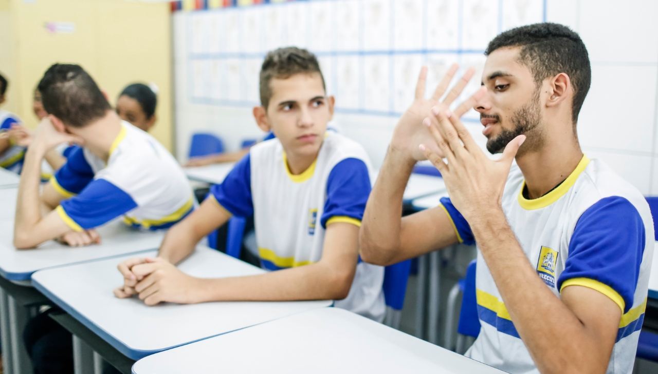 Dois estudantes em sala de aula. Um deles usa Libras enquanto o outro observa. Mais na postagem